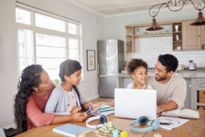 Mixed race family using information technology at home. Two little girls doing homework in the kitchen with help from their mother and father. Parents and children working on school work together