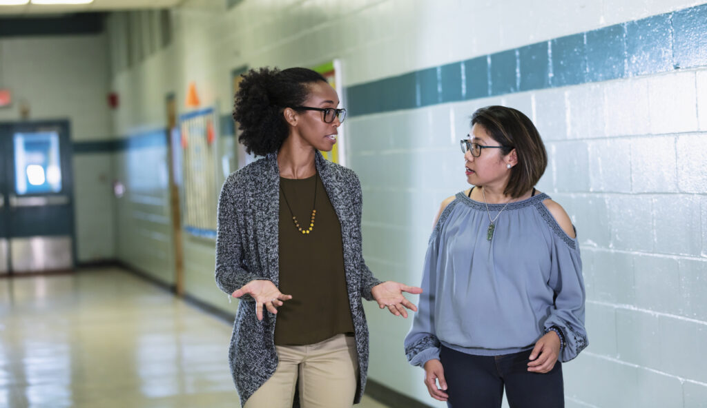 Two multiracial teachers walking and talking in a school hallway. The woman on the left is mixed race Hispanic and African-American, in her 30s. Her coworker is a Filipino woman in her 40s.