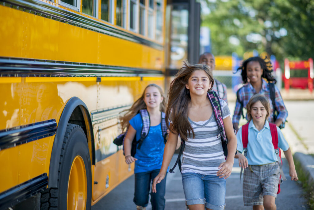 A multi-ethnic group of elementary age children are running from the bus to class. They are smiling and looking at the camera.