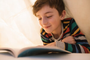 Happy boy reading emotionally book is lying in bed under yellow blanket. Child looks homework into book, reads, running his finger page. Distance learning. Home school education. Quarantine