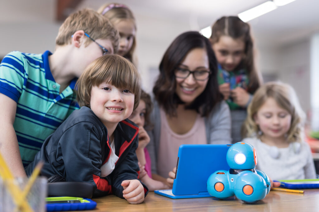 An ethnic female teacher is surrounded by her students as she shows them how to program a robot using a digital tablet. One of the boys is smiling at the camera.