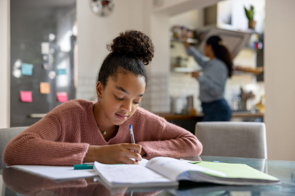 Teenage girl doing her homework while her mother is cooking at the background in the kitchen - lifestyle concepts